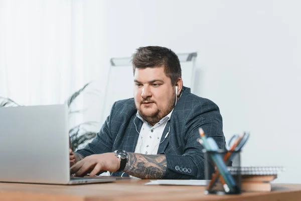 Hombre de negocios con sobrepeso escuchando música con auriculares mientras trabaja en el ordenador portátil en la oficina - foto de stock