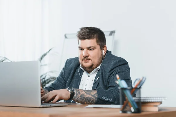 Homme d'affaires en surpoids écouter de la musique avec des écouteurs tout en travaillant sur un ordinateur portable au bureau — Photo de stock