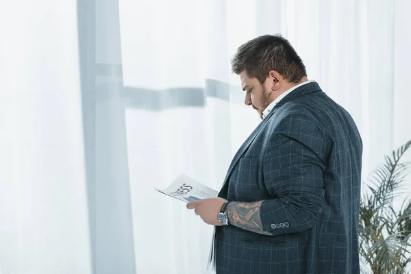 Overweight businessman in suit reading newspaper at window — Stock Photo