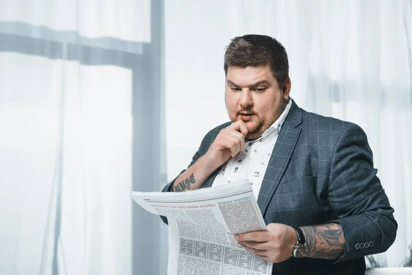 Thoughtful overweight businessman in suit reading newspaper in office — Stock Photo