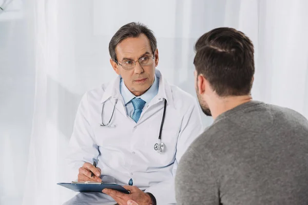 Médico sujetando el portapapeles y escuchando paciente - foto de stock