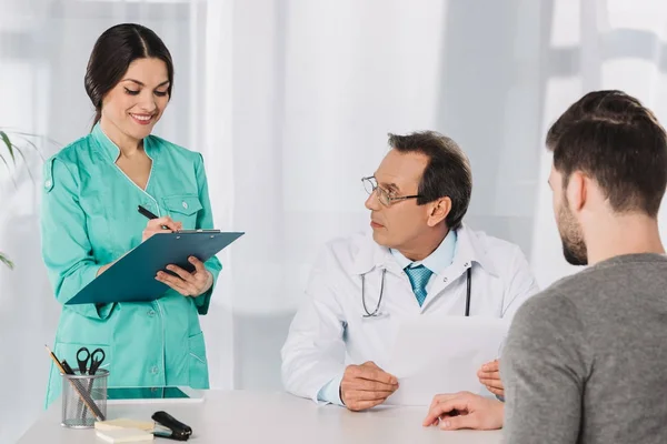 Doctor mirando sonriente enfermera escribiendo algo a portapapeles - foto de stock