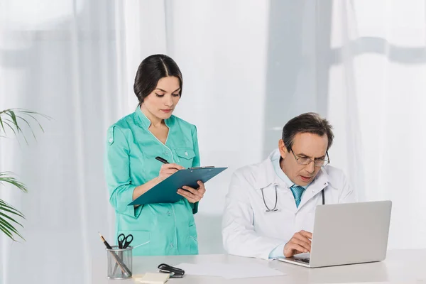 Nurse writing to clipboard and doctor using laptop — Stock Photo