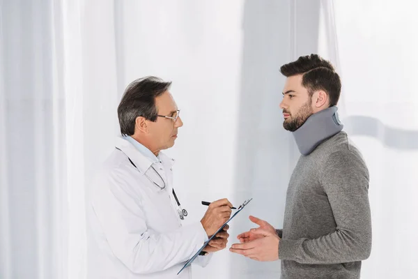 Doctor writing down to clipboard after patient in neck brace — Stock Photo