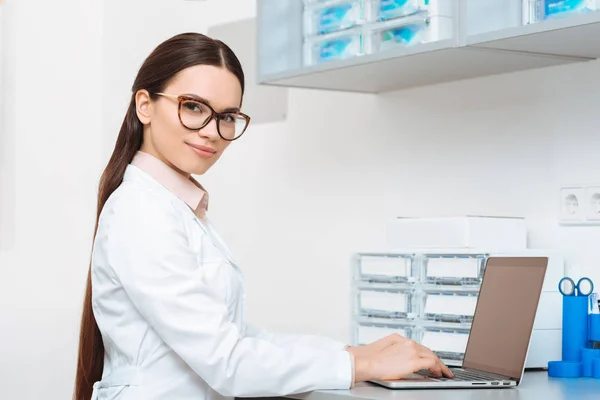 Side view of smiling doctor in white coat looking at camera while working on laptop in clinic — Stock Photo