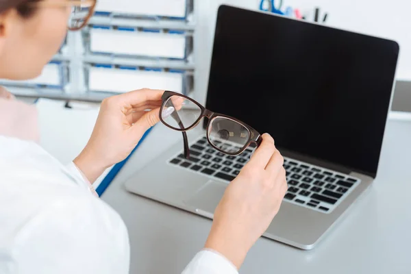 Foyer sélectif de l'ophtalmologiste avec des lunettes dans les mains et ordinateur portable sur la table — Photo de stock