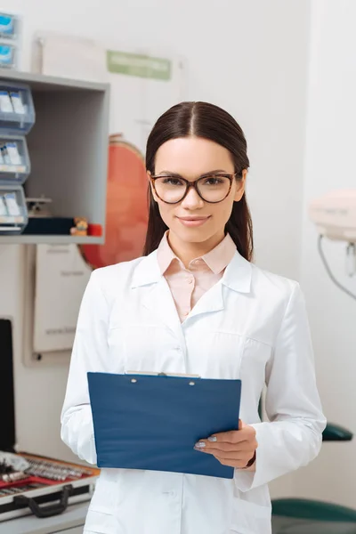 Portrait of smiling optometrist in white coat with notepad in hands looking at camera — Stock Photo