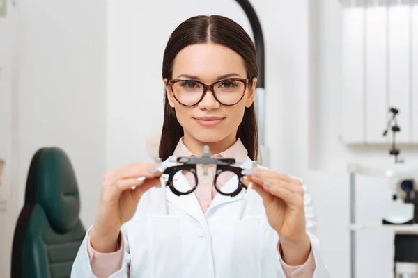 Portrait of young optometrist in white coat showing trial frame in hands in clinic — Stock Photo