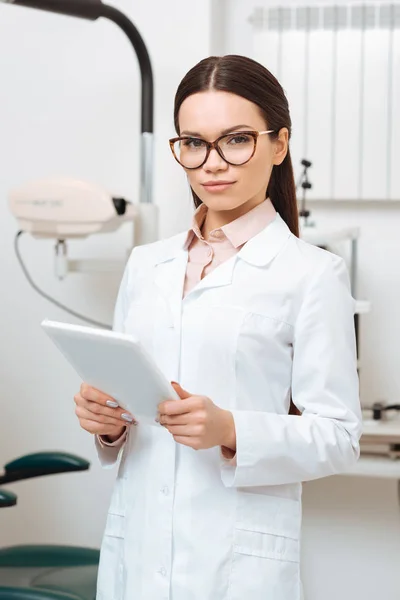 Retrato de joven optometrista con tableta digital en las manos mirando a la cámara en la clínica - foto de stock