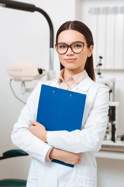 Portrait of smiling optometrist in white coat with notepad in hands looking at camera in clinic — Stock Photo