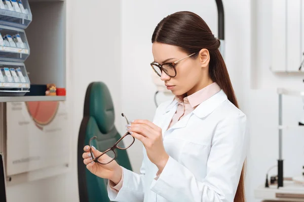 Vue latérale de l'ophtalmologiste focalisé regardant la paire de lunettes dans les mains en clinique — Photo de stock