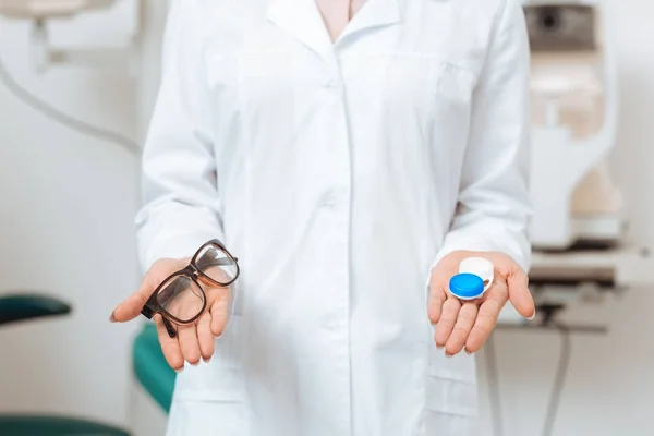 Vista recortada del optometrista en capa blanca sosteniendo gafas y lentes de contacto en la clínica — Stock Photo