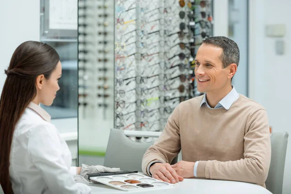 Side view of smiling man looking at oculist while sitting at table in optics — Stock Photo