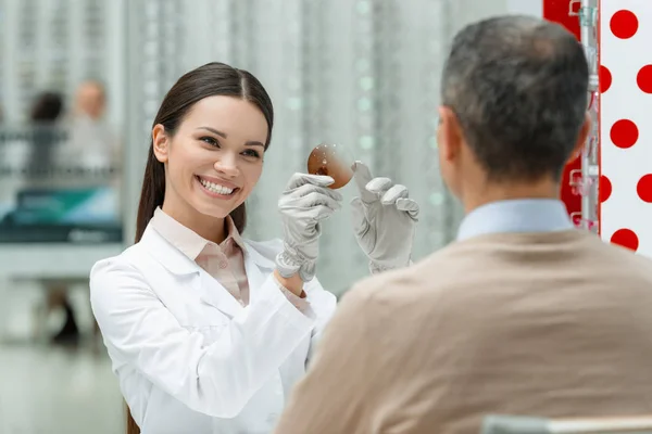 Smiling ophthalmologist in white coat showing lens to patient in optics — Stock Photo