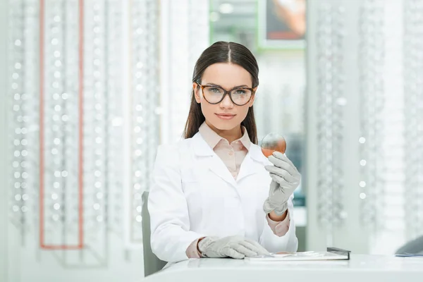 Portrait of oculist in eyeglasses with lens in hand looking at camera in optics — Stock Photo