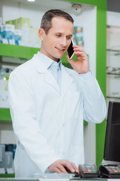 Портрет улыбающегося оптометриста в белом халате разговаривающего на смартфоне у прилавка в оптике — стоковое фото