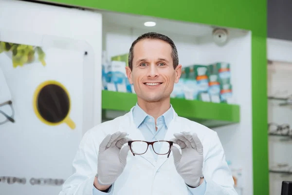 Портрет улыбающегося оптометриста, показывающего очки в руках в оптике — стоковое фото