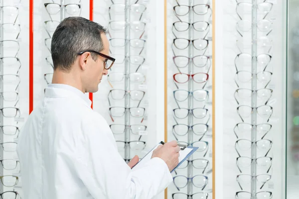 Visão lateral do optometrista fazendo anotações em pé nas prateleiras com óculos em óptica — Fotografia de Stock