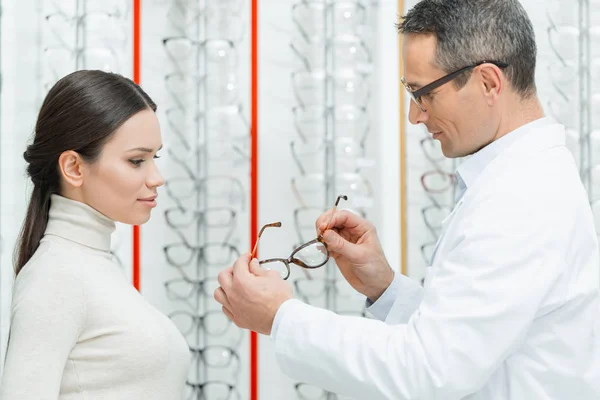 Vista lateral del oculista ayudando a la mujer a elegir un par de gafas en óptica - foto de stock