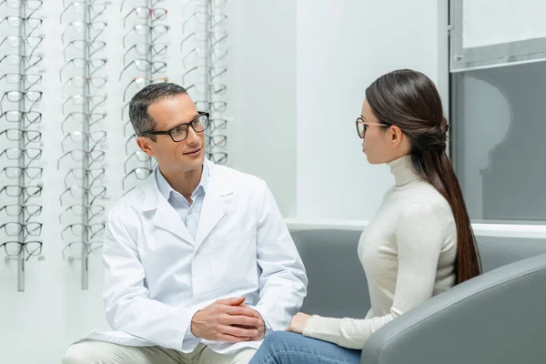 Vista lateral de oftalmólogo y mujer joven conversando en óptica - foto de stock
