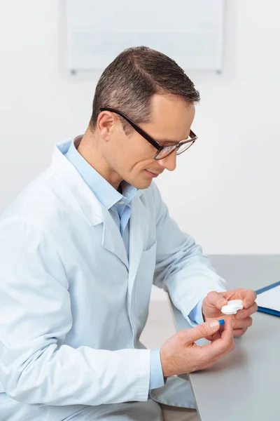 Vista lateral del oftalmólogo mirando la lente de contacto en las manos mientras está sentado en el lugar de trabajo en la clínica - foto de stock