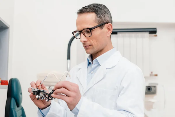 Optometrista in camice bianco guardando cornice di prova in mano in clinica — Foto stock