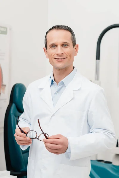 Portrait of smiling doctor in white coat with eyeglasses in hands looking at camera in clinic — Stock Photo