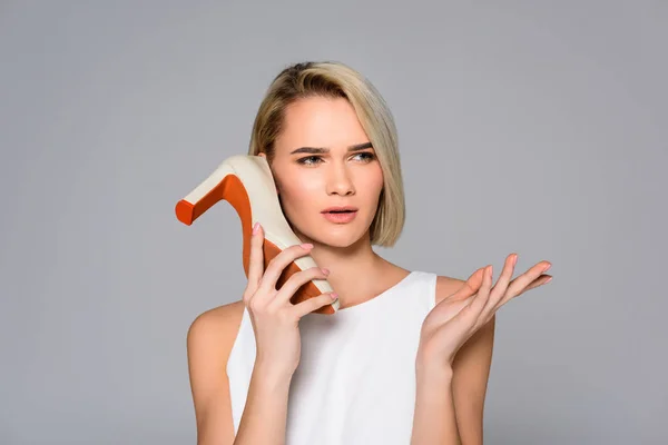 Beautiful confused girl holding heeled shoe as phone, isolated on grey — Stock Photo