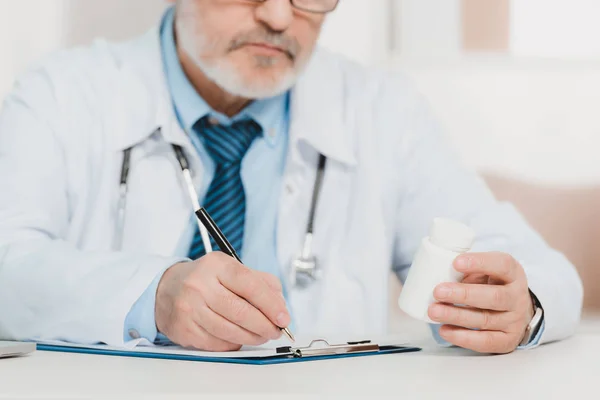 Partial view of doctor with pills in hand making notes on notepad at workplace — Stock Photo