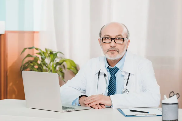 Ritratto del dottore anziano in occhiali che guarda la macchina fotografica mentre si siede sul posto di lavoro con il computer portatile in clinica — Foto stock