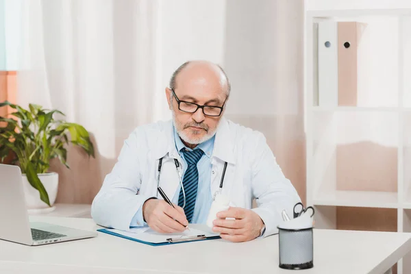 Portrait d'un médecin principal concentré prenant des notes sur un bloc-notes au travail avec un ordinateur portable à la clinique — Photo de stock