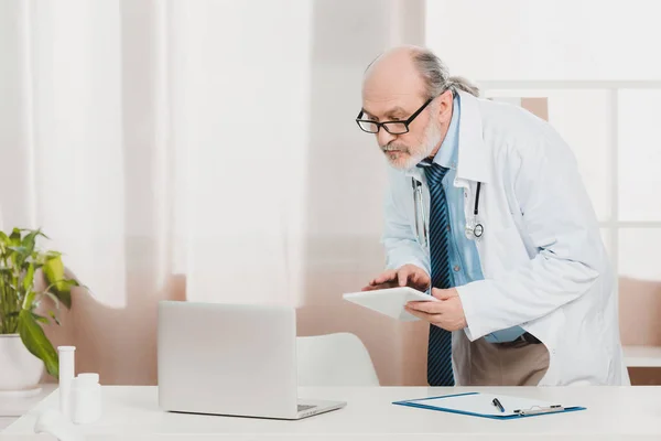 Portrait of senior doctor in white coat with tablet in hands at workplace in clinic — Stock Photo