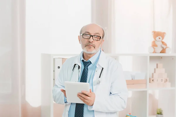 Portrait du médecin aîné en manteau blanc avec tablette dans les mains regardant la caméra à la clinique — Photo de stock