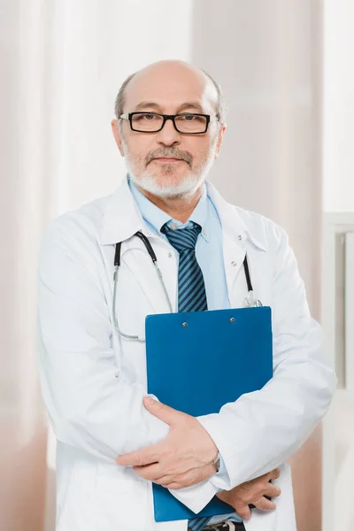 Retrato de médico senior en gafas con estetoscopio y bloc de notas mirando a la cámara - foto de stock