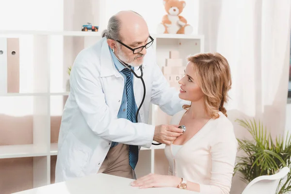 Senior doctor in white coat examining woman with stethoscope in clinic — Stock Photo
