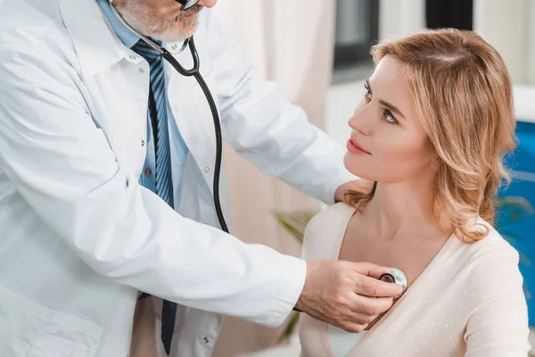 Senior doctor in white coat examining woman with stethoscope in clinic — Stock Photo