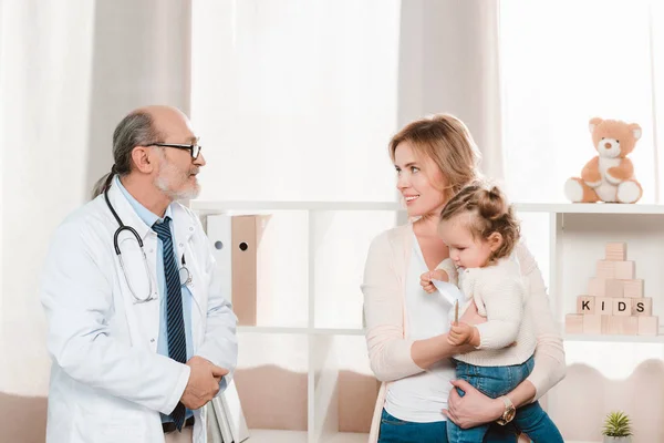 Портрет старшого лікаря, усміхнена мати з маленькою дівчинкою на руках у клініці — стокове фото