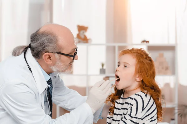 Старший врач в медицинских перчатках осматривает горло маленьких пациентов в клинике — стоковое фото
