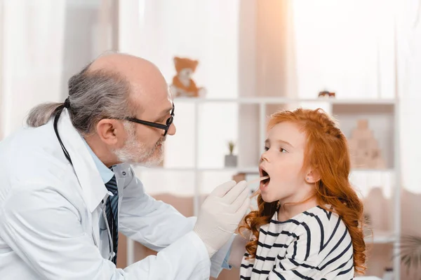 Médico senior en guantes médicos examinando pequeños pacientes garganta en la clínica — Stock Photo