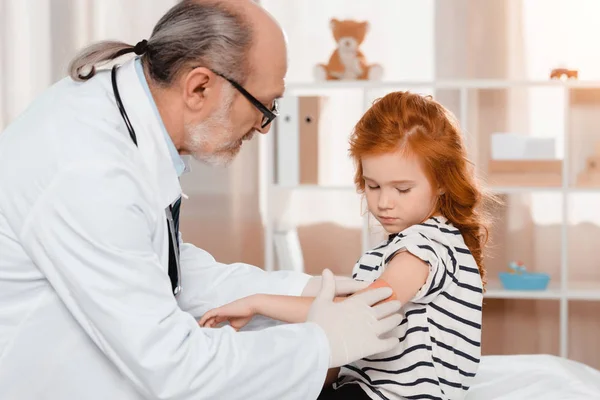 Senior pediatrician putting plaster on patients arm after injection in clinic — Stock Photo