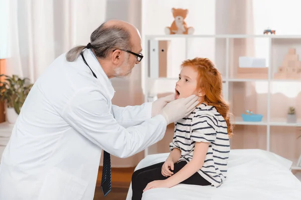 Médico senior en guantes médicos examinando pequeños pacientes garganta en la clínica - foto de stock