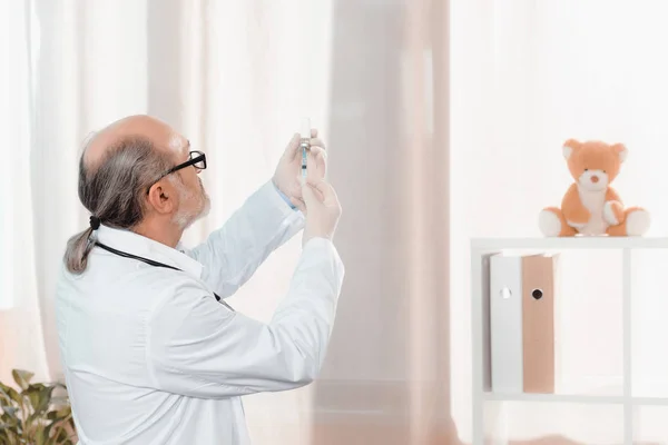 Вид со стороны старшего врача в очках и медицинских перчатках, держащего шприц в клинике — стоковое фото