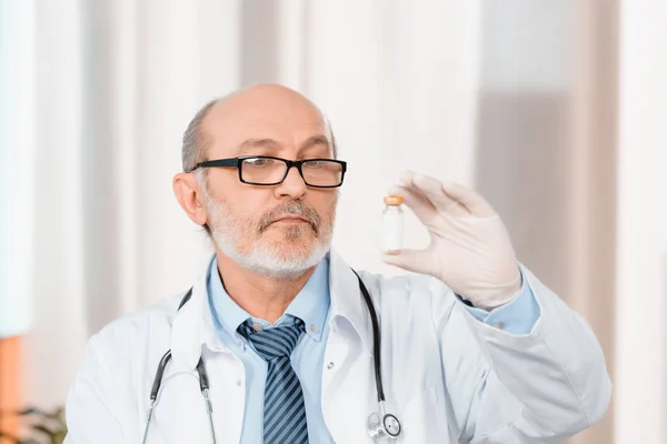 Retrato de médico senior en anteojos y guantes médicos mirando medicamentos en la mano en la clínica - foto de stock