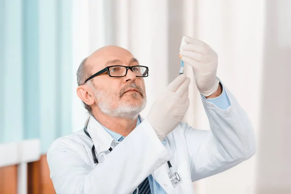 Portrait of senior doctor in eyeglasses and medical gloves holding syringe in clinic — Stock Photo
