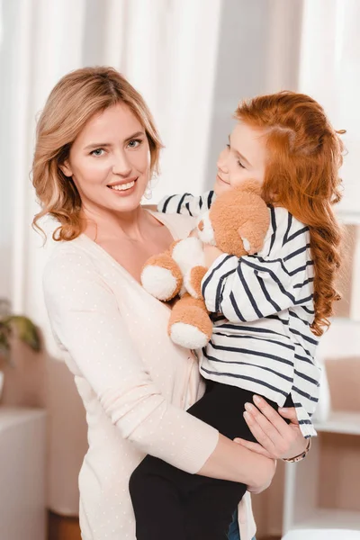 Retrato de madre sonriente sosteniendo a la pequeña hija con oso de peluche - foto de stock