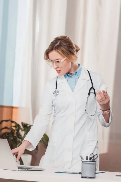 Retrato del médico de bata blanca con medicamentos en la mano usando portátil en el lugar de trabajo en la clínica - foto de stock