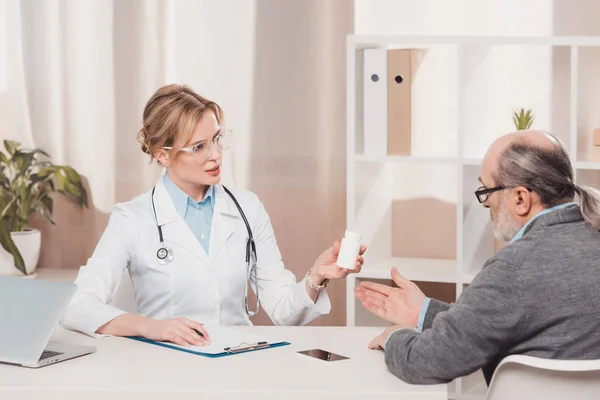 Doctor in white coat and eyeglasses showing medicines to senior patient at workplace in clinic — Stock Photo