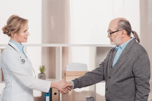 Seitenansicht eines lächelnden Arztes im weißen Kittel und Patienten beim Händeschütteln in der Klinik — Stockfoto