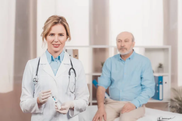 Selective focus of doctor holding syringe for injection with senior patient behind in clinic — Stock Photo