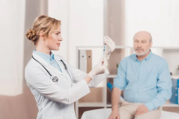 Selective focus of doctor holding syringe for injection with senior patient behind in clinic — Stock Photo